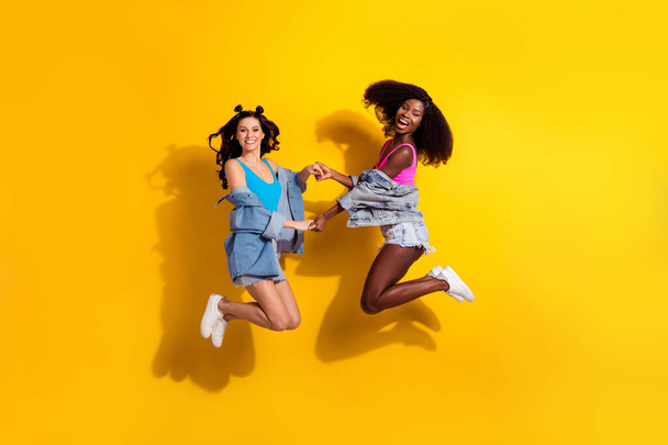 Photo of two adorable pretty young ladies wear jeans outfit holding arms jumping arms isolated yellow color background - Фото, изображение