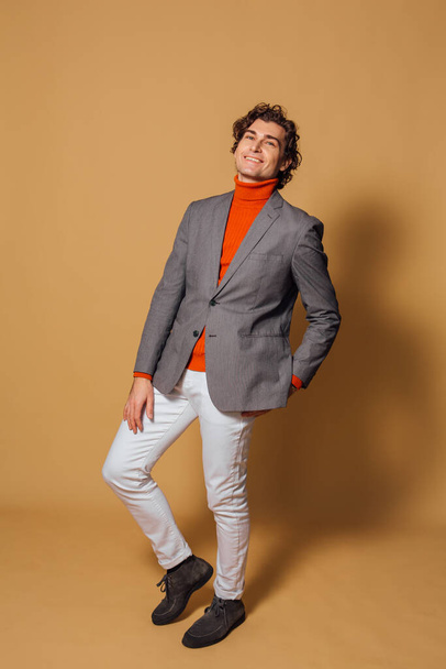 Fashion portrait of a tall handsome man dressed in orange turtleneck, white jeans and grey jacket posing on the beige background - Фото, изображение