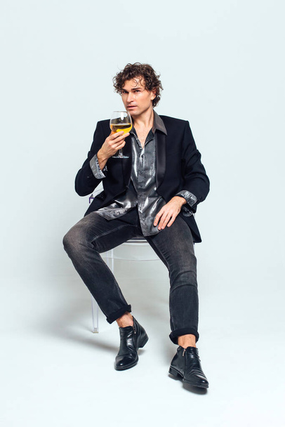Tall handsome confident young man in a jacket sitting on the chair and holding glass of wine over white background - Zdjęcie, obraz