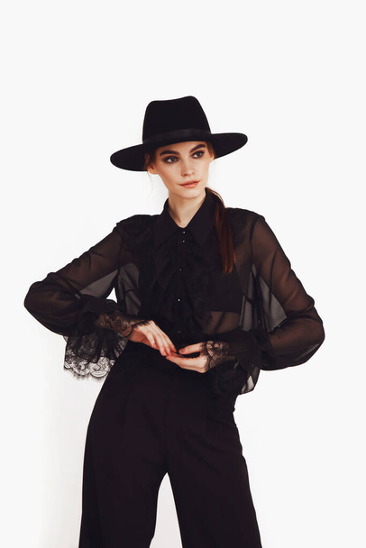 Stylish cute young businesswoman in black elegant blouse and hat on white light background. Woman in fashionable clothes posing at camera. Concept of style, fashion, beauty and achievement of goals - 写真・画像