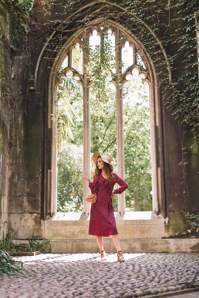 Stylish woman in a hat and polka dot dress. Beauty. Polka dot dress. Burgundy in polka dots. In full growth. Background architecture. Gothic architectures. - Foto, immagini