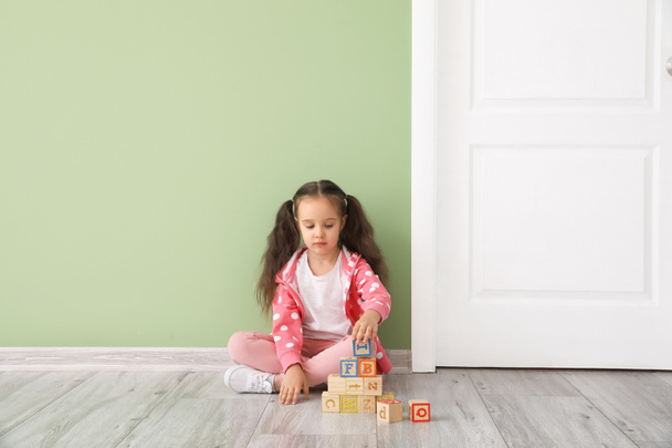 Маленькая девочка с деревянными кубиками сидит возле закрытой двери - Фото, изображение