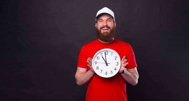 Fotografie usmívajícího se mladého muže s plnovousem v červeném tričku zobrazující velké nástěnné hodinky na černém pozadí. - Fotografie, Obrázek