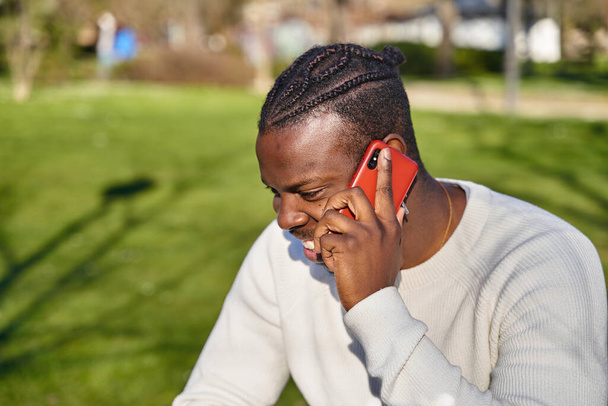 Hombre negro feliz hablando por teléfono inteligente sentado en un parque en un día soleado.  - Foto, Imagen
