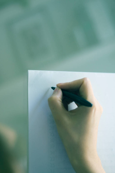 Νεαρή κυρία που γράφει σε χαρτί με μολύβι στο χέρι στο τραπέζι. - Φωτογραφία, εικόνα