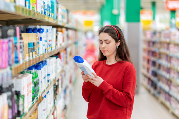 A young Caucasian woman in a red sweater holds a bottle of shampoo and reads the ingredients on the label. In the background, shelves with cosmetics. The concept of buying cosmetics. - Foto, Imagem