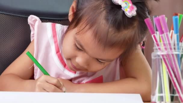 Милая маленькая девочка рисует радугу на бумаге с цветными карандашами за столом дома. - Кадры, видео