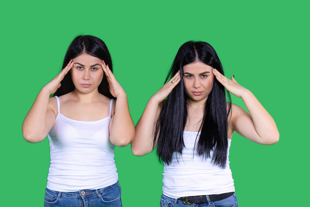 Young twin girls, business women pointing fingers to foreheads on the way of stressful, serious thinking, migraine headache. Fashion brunette, caucasian style on green background. High quality photo - Photo, image