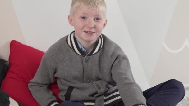 Un chico rubio lindo de 10 años se sienta en el sofá y sonríe - Imágenes, Vídeo