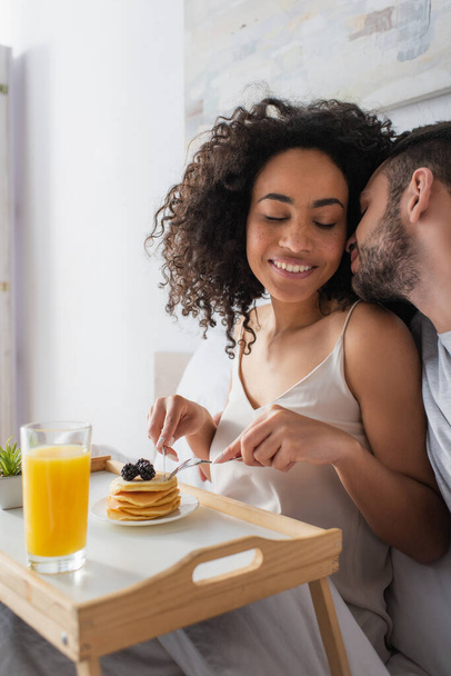 bearded man kissing happy african american woman holding cutlery near pancakes in bedroom - Zdjęcie, obraz