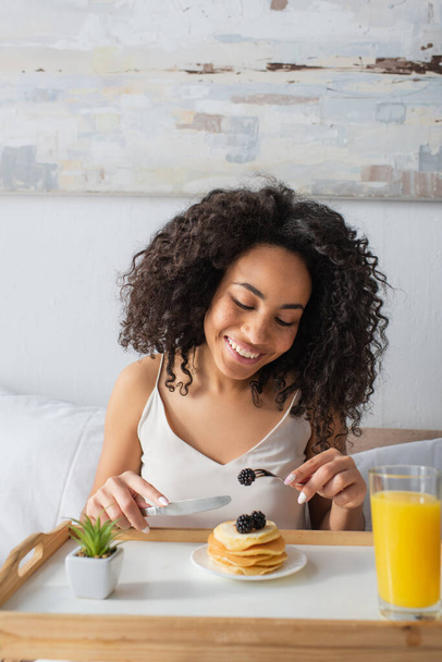 happy african american woman holding fork with blackberry near pancakes on tray - Photo, Image