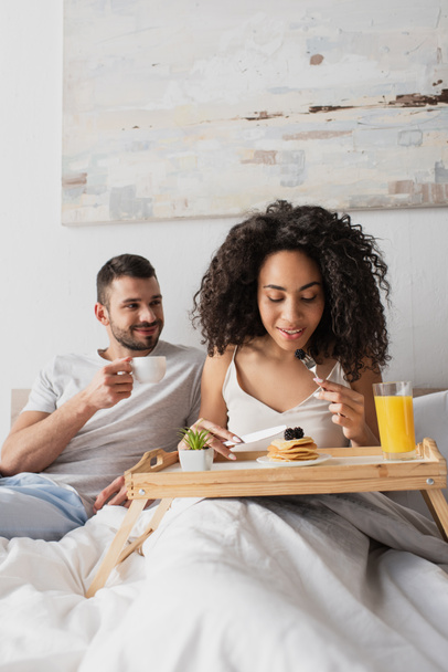 happy african american woman holding fork with blackberry near pancakes on tray and boyfriend with cup of coffee - Fotoğraf, Görsel