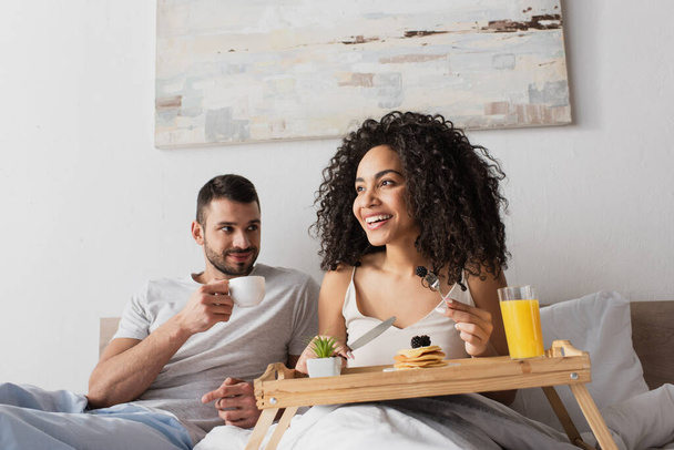 smiling african american woman holding fork with blackberry near pancakes on tray and boyfriend with cup of coffee - Foto, immagini