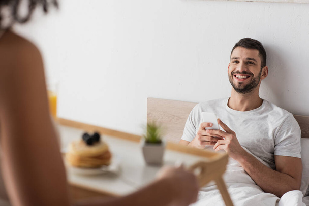 hombre feliz con teléfono inteligente mirando a la novia afroamericana sosteniendo bandeja con desayuno en primer plano borrosa  - Foto, Imagen