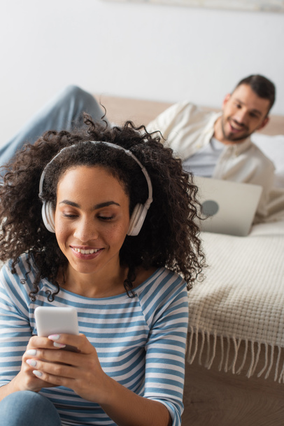 smiling african american woman in wireless headphones holding smartphone near boyfriend using laptop on blurred background - Zdjęcie, obraz
