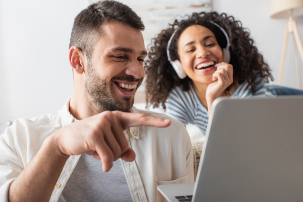 happy man pointing with finger at laptop near african american woman in wireless headphones on blurred background - Fotó, kép