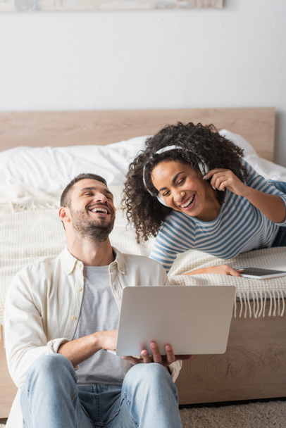 interracial couple laughing near gadgets in bedroom - Photo, Image