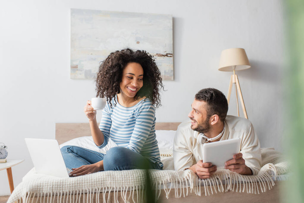 happy african american woman sitting on bed, holding cup and using laptop near boyfriend with digital tablet  - Φωτογραφία, εικόνα