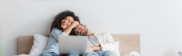 cheerful interracial couple resting in bed near laptop, banner - Fotoğraf, Görsel