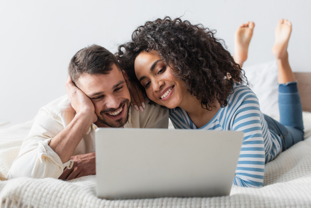 joyful interracial couple chilling on bed while watching movie on laptop  - Foto, Imagem