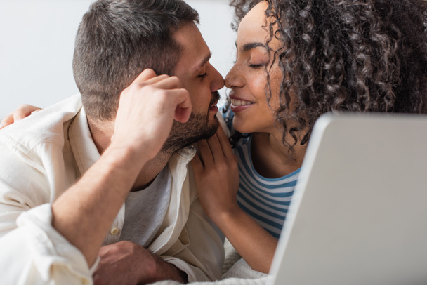 interracial couple kissing while chilling on bed near laptop  - Zdjęcie, obraz