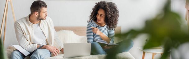 african american woman pointing at boyfriend sitting with folder near laptop on bed, banner - Foto, afbeelding