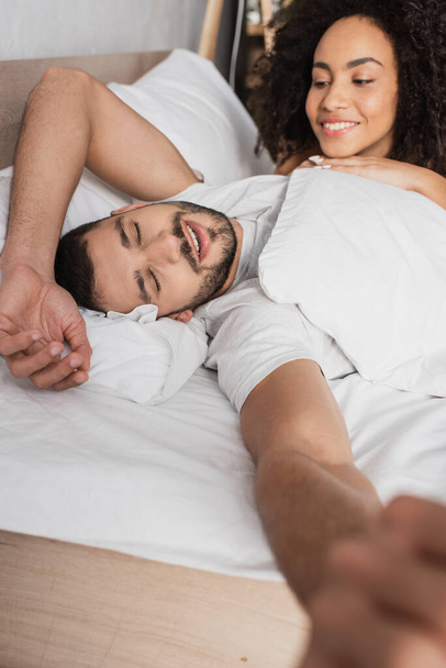 happy african american woman looking at sleepy boyfriend in bed - Foto, afbeelding