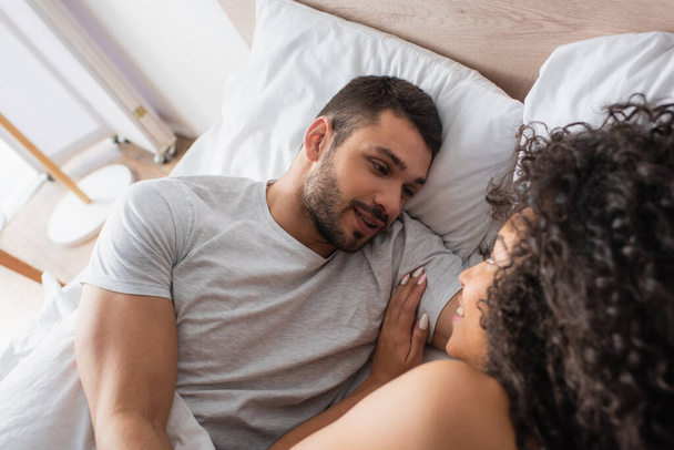 high angle view of positive interracial couple looking at each other while lying in bed at home - Foto, afbeelding