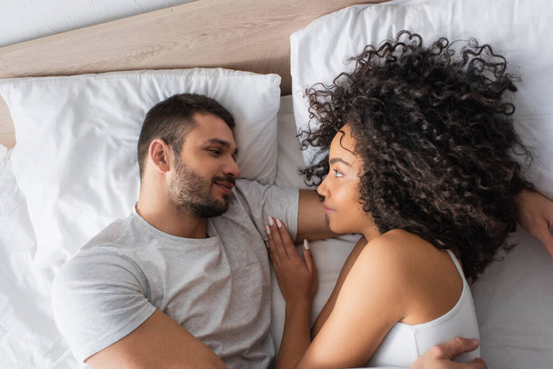 top view of pleased interracial couple looking at each other while lying in bed at home - Fotoğraf, Görsel