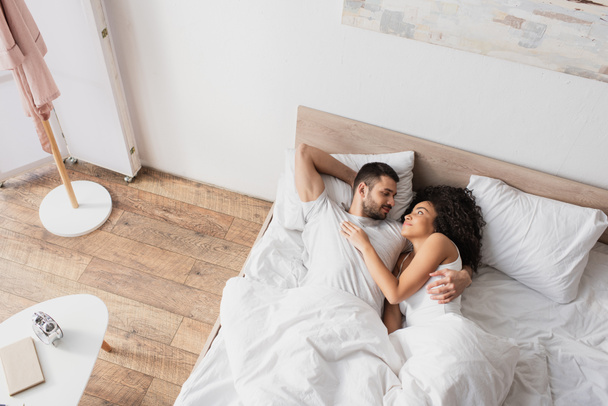vista superior de pareja interracial positiva abrazándose y acostado en la cama - Foto, Imagen