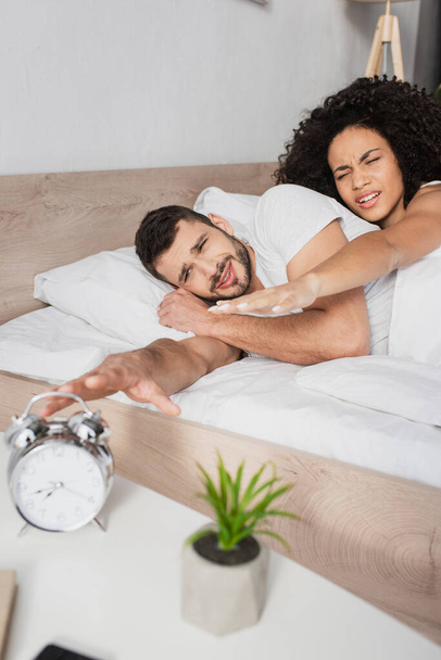 displeased interracial couple reaching alarm clock in morning  - Fotografie, Obrázek