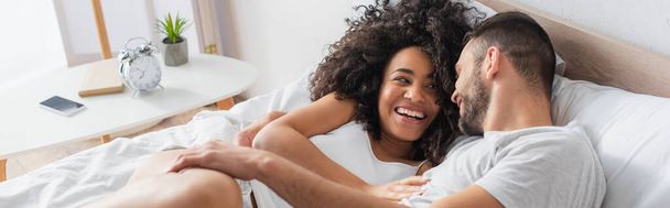 cheerful interracial couple smiling and lying on bed, banner - Photo, Image