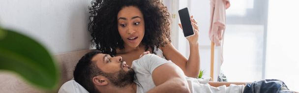 meglepett afro-amerikai nő néz barátja, miközben gazdaság smartphone üres képernyő a hálószobában, banner - Fotó, kép