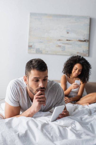 szakállas férfi fekszik az ágyon, és sms-ezés okostelefon közelében afro-amerikai nő elmosódott háttér  - Fotó, kép