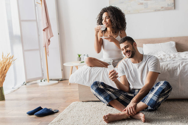 mujer afroamericana feliz sosteniendo taza con café mientras novio barbudo usando teléfono inteligente y sentado en la alfombra - Foto, Imagen