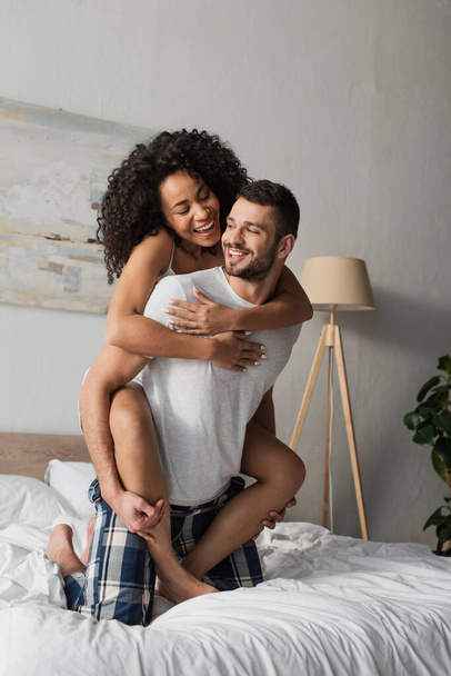 cheerful bearded man piggybacking african american girlfriend in bedroom - Photo, Image