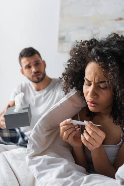 sick african american woman looking at digital thermometer near worried boyfriend on blurred background  - Foto, afbeelding