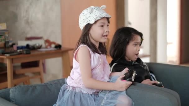 Twee Aziatische meisjes zitten op een grijze fauteuil en genieten van videospelletjes in hun woonkamer. - Video