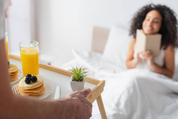vue recadrée de l'homme tenant plateau avec petit déjeuner savoureux près heureuse petite amie afro-américaine sur fond flou - Photo, image