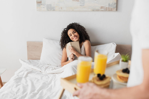 hombre borroso sosteniendo bandeja con desayuno cerca de la novia afroamericana feliz con libro en la cama - Foto, Imagen