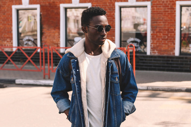 Portrait of stylish young african american man model wearing denim jacket with backpack posing on a city street - Фото, изображение