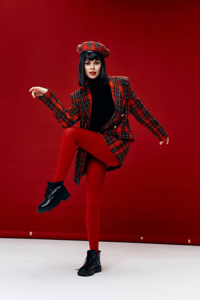 woman raised her leg up on a red background plaid coat hat gesturing with her hands - Photo, Image