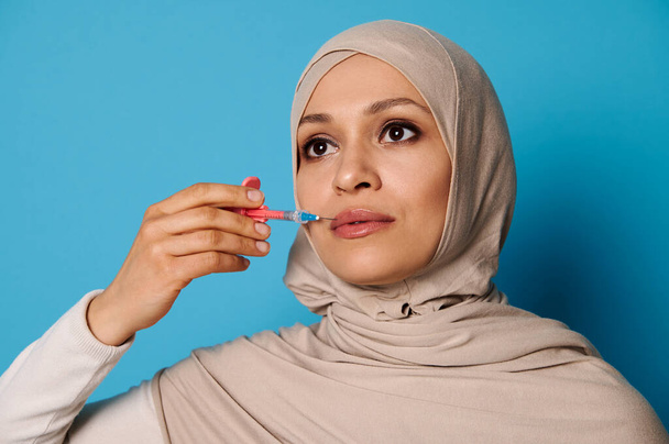 Primo piano della bella donna musulmana in hijab che tiene una siringa con iniezione di bellezza vicino alle labbra. Concetto di aumento delle labbra in cosmetologia per iniezione. Copia spazio, sfondo blu - Foto, immagini