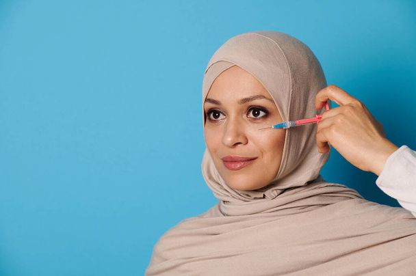 Приваблива молода мусульманка з ідеальною косметикою і чистою здоровою шкірою, поставляючи шприц біля її обличчя на синьому фоні. Простір для тексту - Фото, зображення