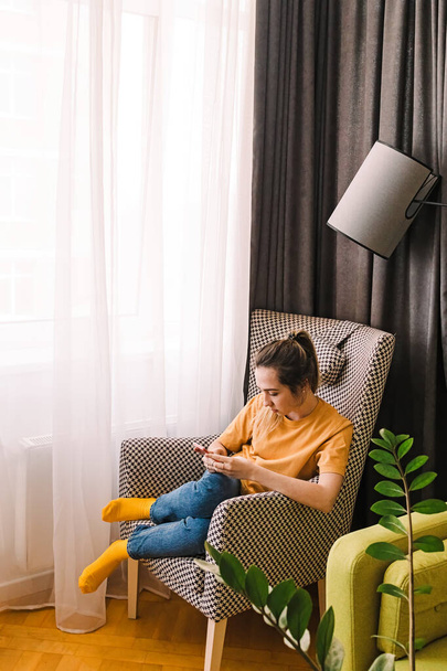 Young woman with smartphone sitting in the comfortable armchair at home - 写真・画像