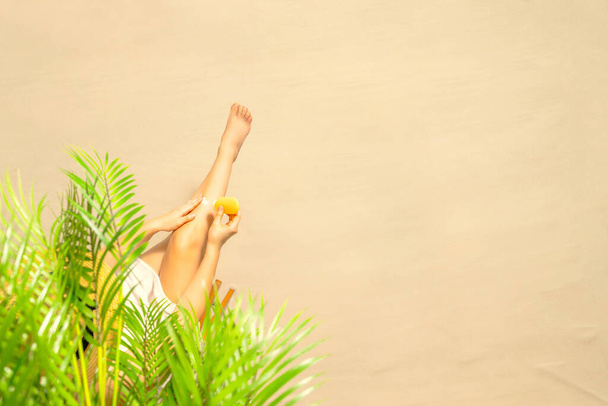 Alone woman applying sunscreen on skin sitting under palm tree branches. Female relaxation on the sand of the beach at summer vacation. Top view - 写真・画像