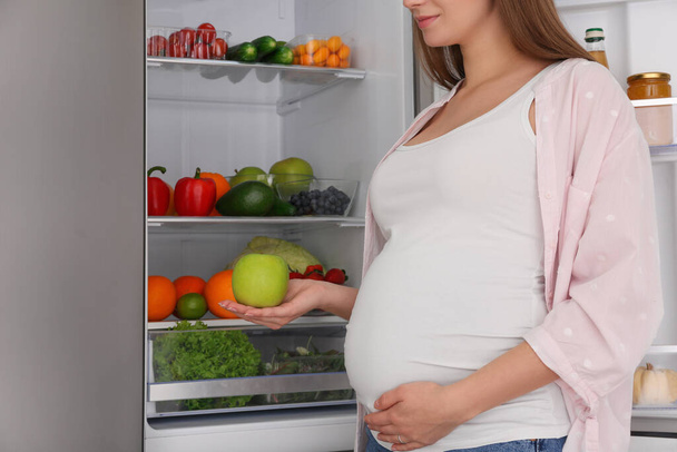Young pregnant woman with apple near fridge at home, closeup. Healthy eating - Foto, afbeelding