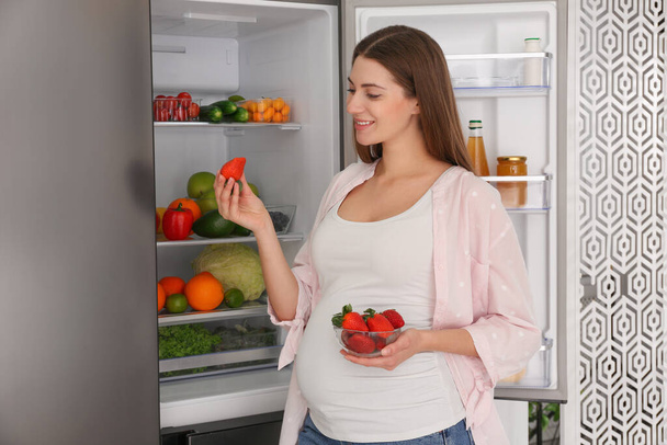 Junge schwangere Frau mit Erdbeeren in der Nähe des Kühlschranks zu Hause. Gesunde Ernährung - Foto, Bild