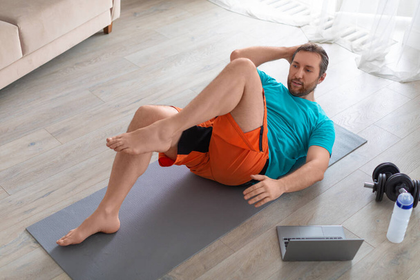 Trening online w domu. Mężczyzna wykonuje ćwiczenia brzuszne przed monitorem laptopa. Trenuje z internetowym przewodnikiem. - Zdjęcie, obraz