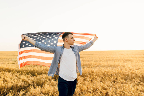 Young man holding American flag while standing on wheat field against blue sky background - Fotó, kép
